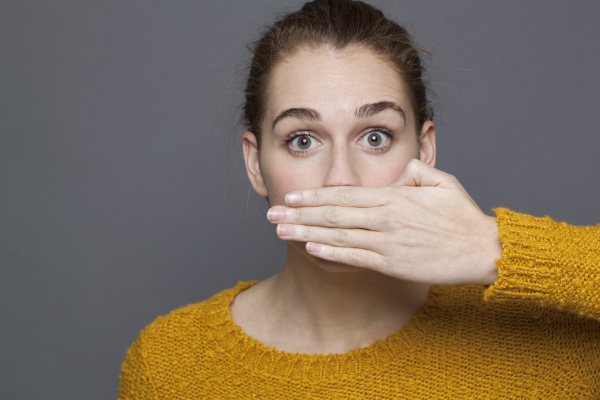 brunette woman in yellow sweater hand in front of mouth looking worried about bad breath in front of gray background