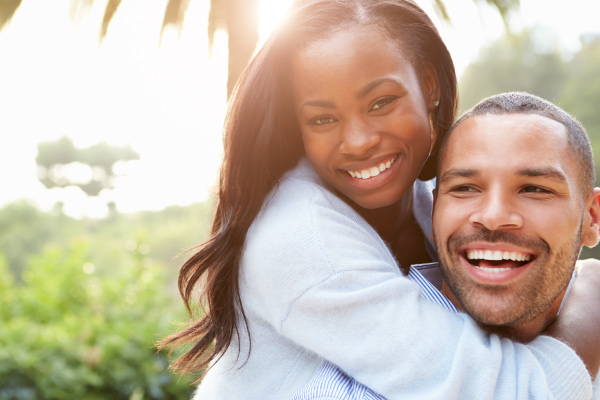 A brunette woman embraces a dark-haired man from behind as they show off their professionally whitened smiles