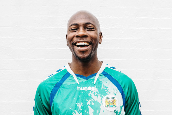 Bald man excited about xylitol wears a teal soccer jersey in front of a white brick wall and smiles with white teeth